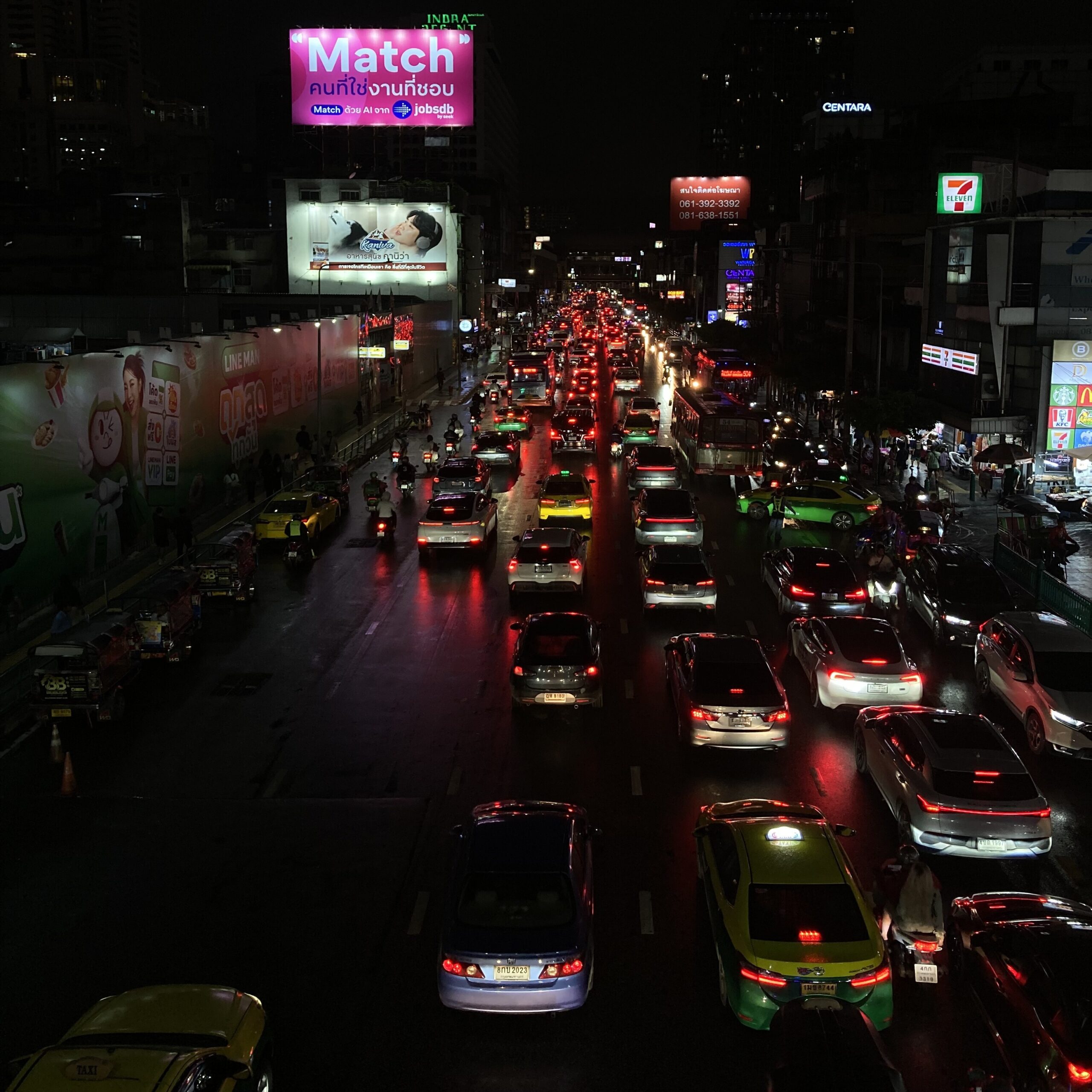 バンコクの夜の大通りに渋滞した車列。ネオンサインが輝く中、タクシーやバイクが行き交う
